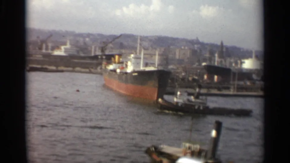1965 Stockholm waterfront with a large freighter, tugboat, harbor cranes, industrial shore structures, and rippled water in the foreground.