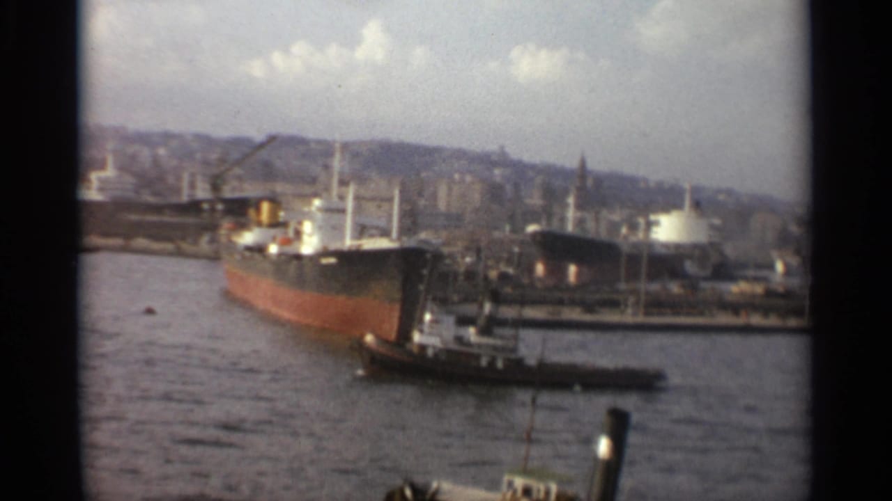 Stockholm harbor home-movie frame from 1965 with a foreground tugboat, cargo ships, dock structures, cranes, and a cloudy skyline.