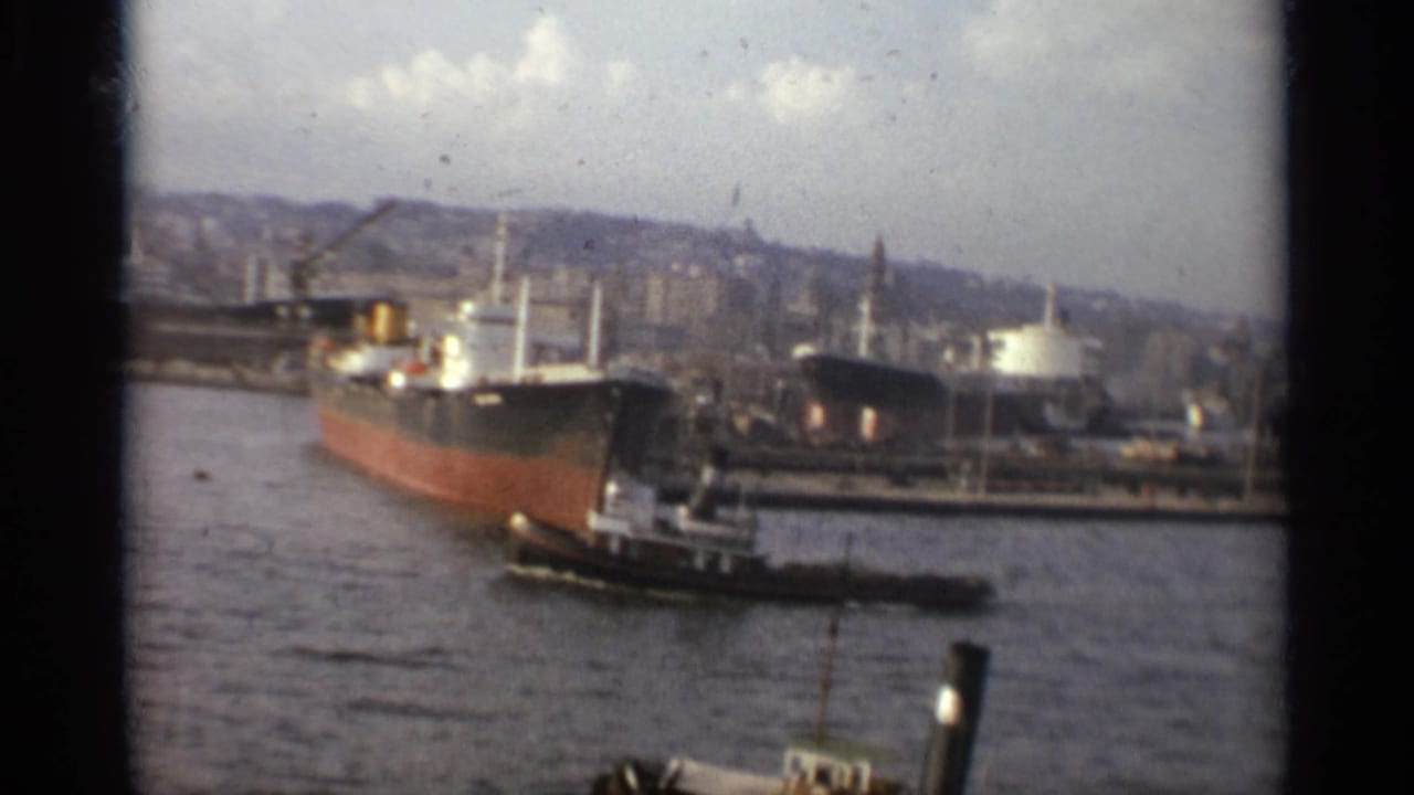 1965 Stockholm harbor view with a docked ocean liner, tugboat, water, and hillside skyline in soft 8mm color.
