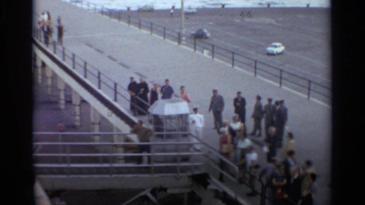 Elevated 1965 Stockholm pier view showing civilians and uniformed figures gathered near railings as cars pass nearby.