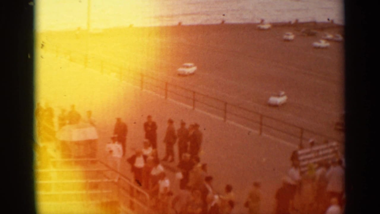 Warmly flared 8mm frame of a Stockholm pier crowd near railings and a waterfront road, with people and cars partly washed by exposure.