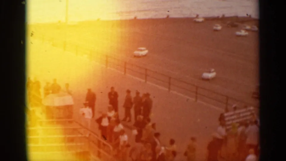 Warmly flared 8mm frame of a Stockholm pier crowd near railings and a waterfront road, with people and cars partly washed by exposure.