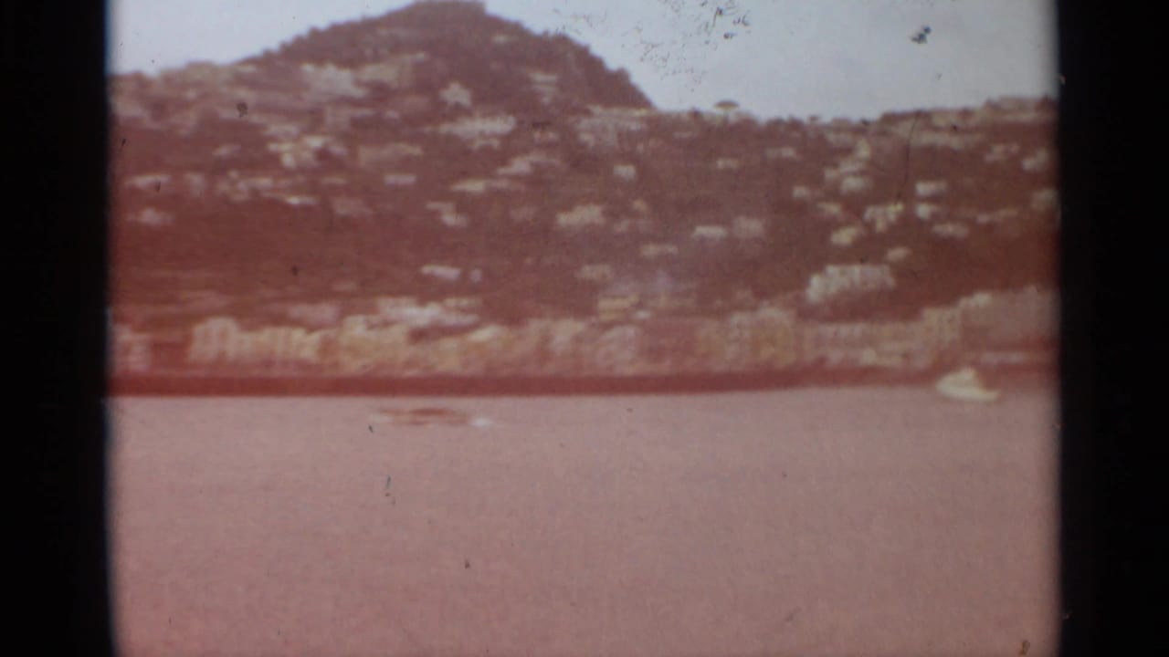 1965 Stockholm home-movie view of calm water below a green hillside with scattered houses and a small white boat near shore.