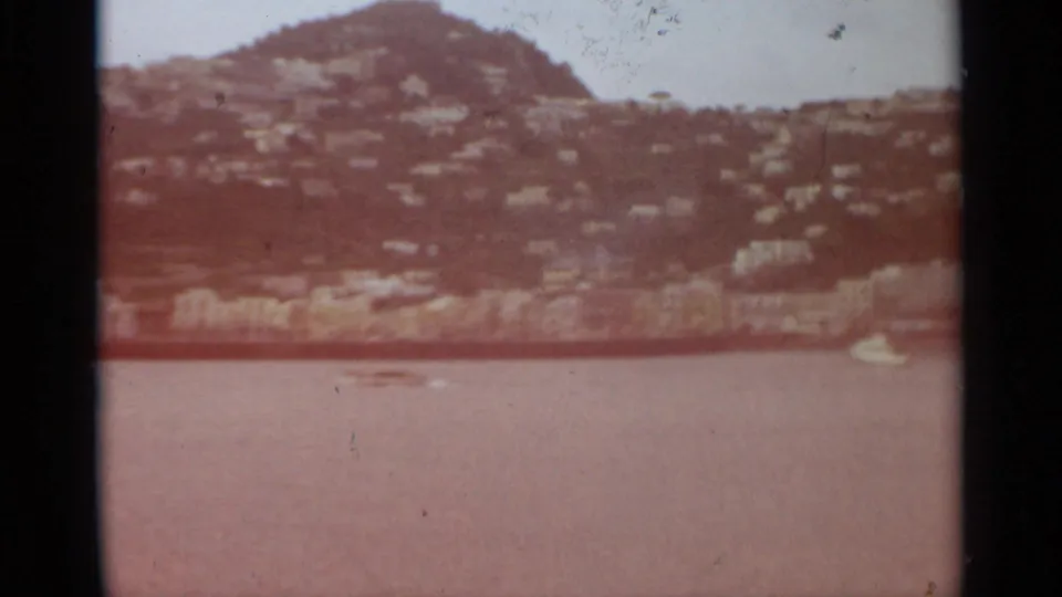 1965 Stockholm home-movie view of calm water below a green hillside with scattered houses and a small white boat near shore.