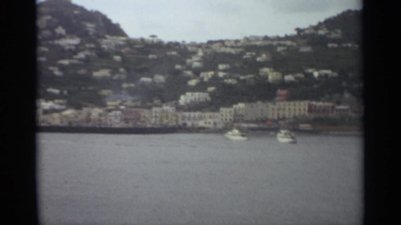Stockholm 1965 8mm frame showing two white boats on water with pale waterfront buildings and houses on the hillside beyond.
