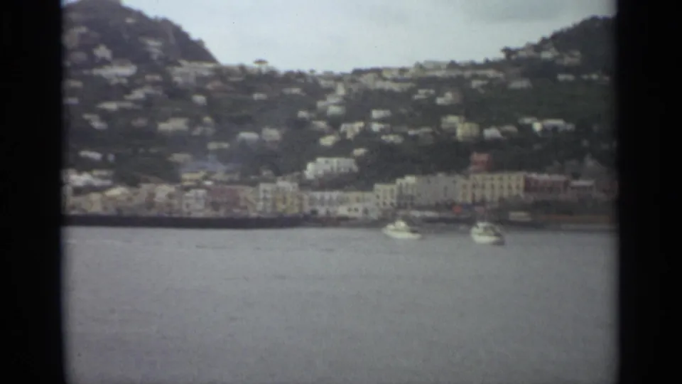 Stockholm 1965 8mm frame showing two white boats on water with pale waterfront buildings and houses on the hillside beyond.