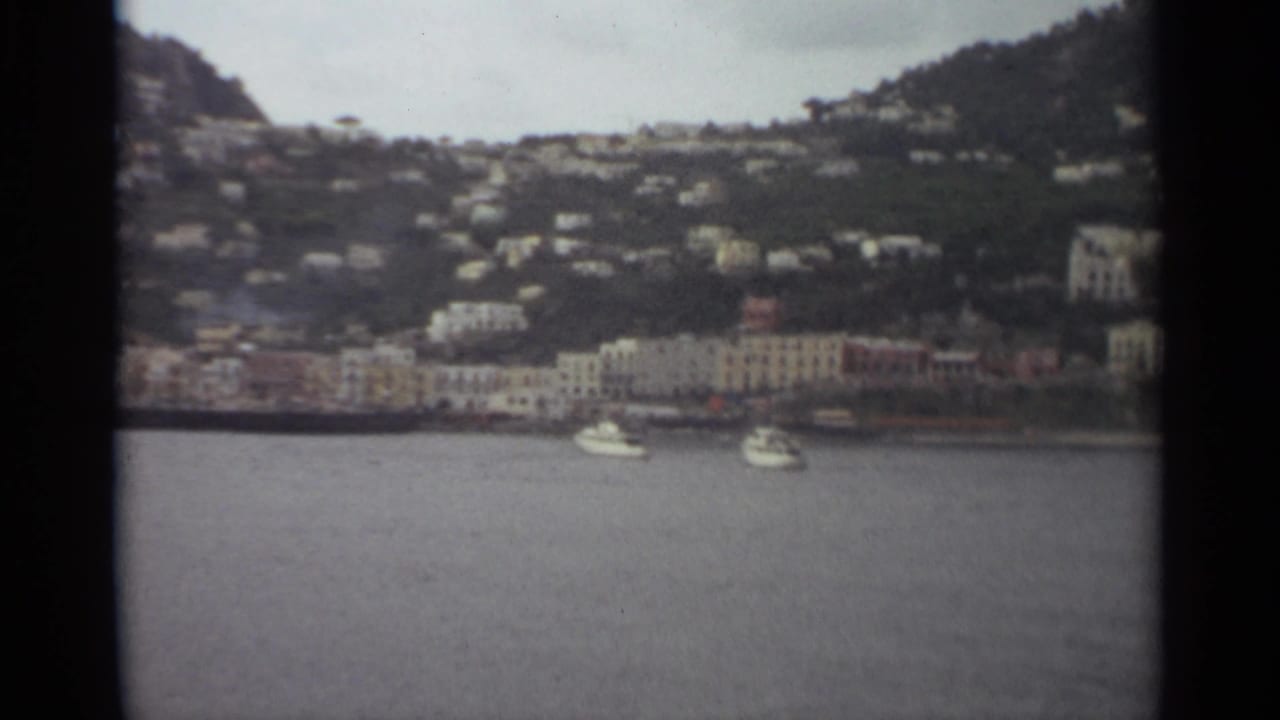 1965 Stockholm home-movie frame with small white boats, a waterfront row of buildings, and houses spread across a green hillside.
