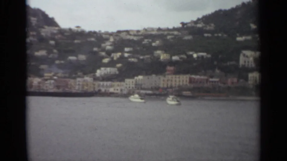 1965 Stockholm home-movie frame with small white boats, a waterfront row of buildings, and houses spread across a green hillside.