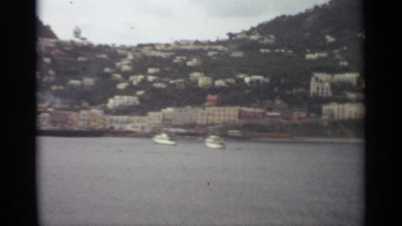 1965 8mm view of a Stockholm waterfront townscape with boats on the water, pale shoreline buildings, and houses rising into green hills.