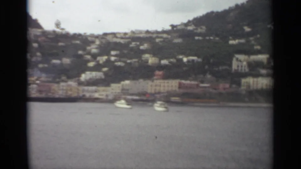 1965 8mm view of a Stockholm waterfront townscape with boats on the water, pale shoreline buildings, and houses rising into green hills.