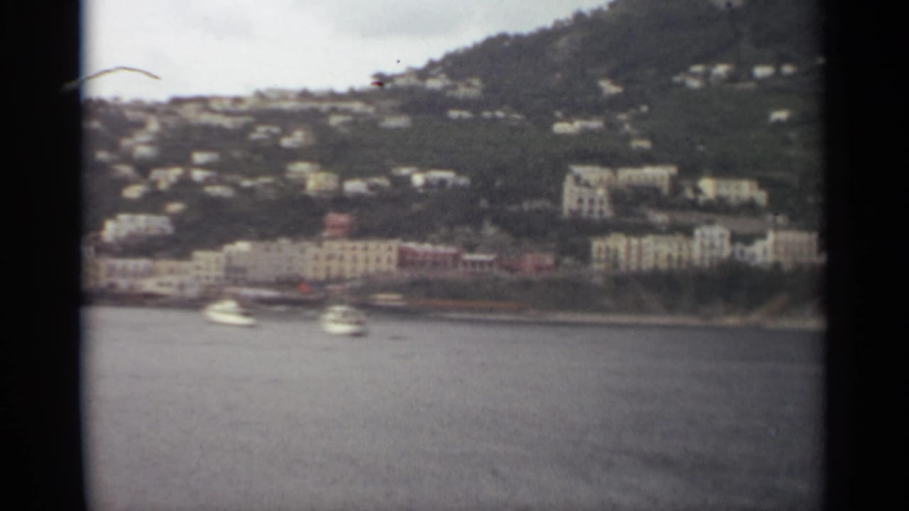 Distant shoreline buildings and boats below a green hillside, framed by dark film edges.