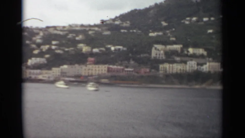 1965 Stockholm home-movie frame showing small boats near a waterfront lined with pale buildings and hillside homes.