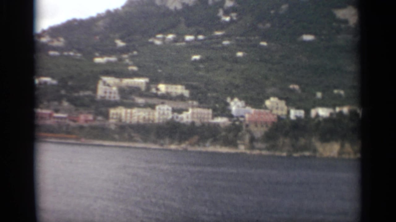 1965 8mm Stockholm waterfront view with calm sea in front, pale buildings along shore, and homes set into a green hillside.