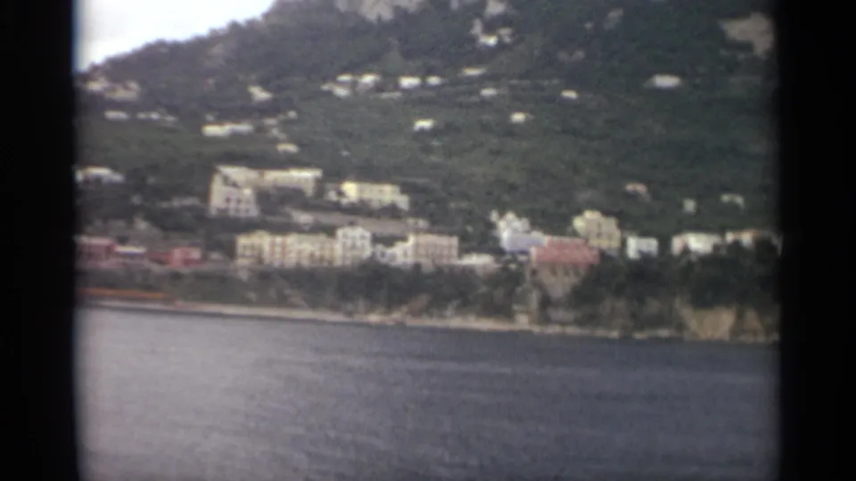 1965 8mm Stockholm waterfront view with calm sea in front, pale buildings along shore, and homes set into a green hillside.