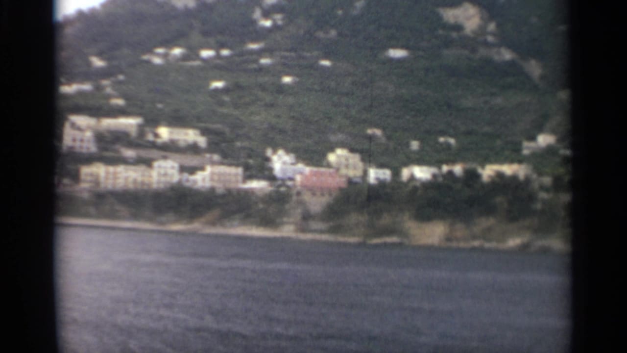 1965 8mm frame showing pale and pastel buildings on a wooded shoreline across calm water, with dark film borders visible.