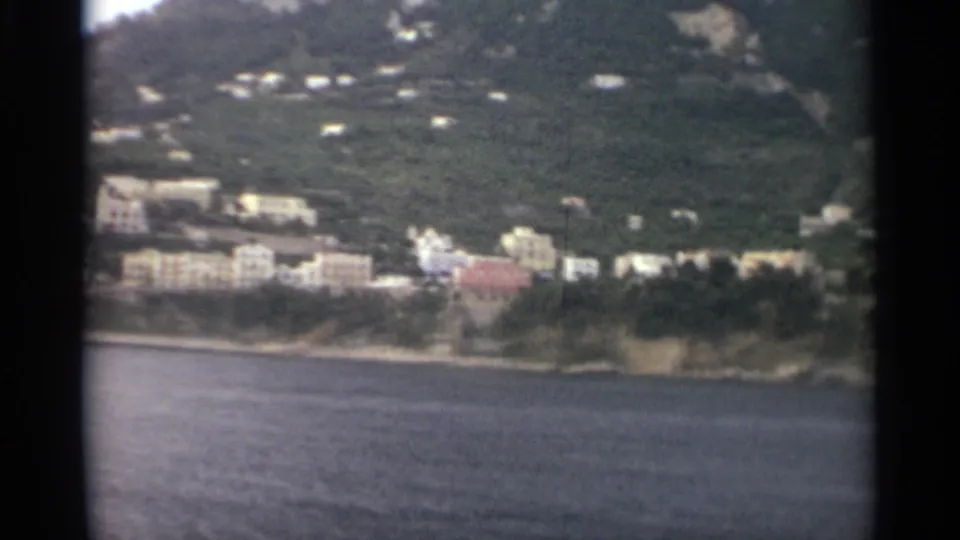 1965 8mm frame showing pale and pastel buildings on a wooded shoreline across calm water, with dark film borders visible.