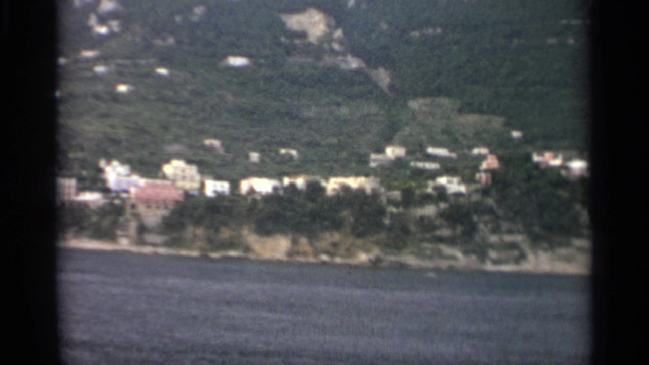 1965 8mm frame with a waterside settlement, trees, and hillside vegetation viewed from across the water during a slow pan.