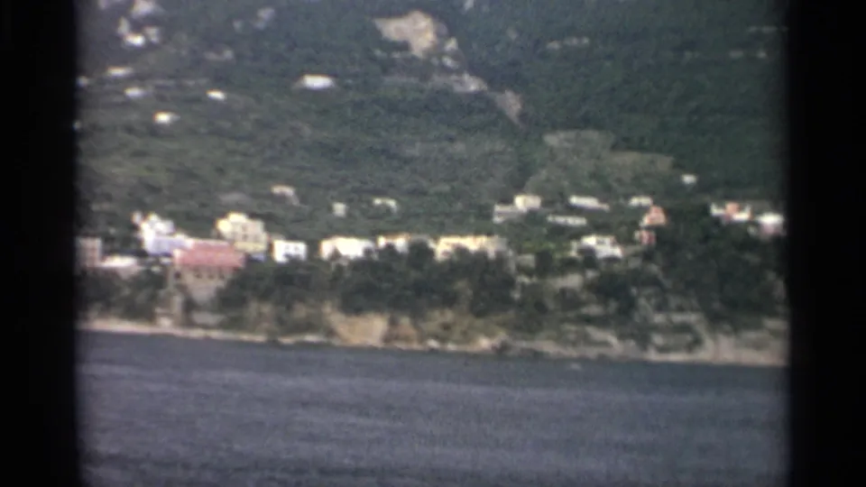 1965 8mm frame with a waterside settlement, trees, and hillside vegetation viewed from across the water during a slow pan.