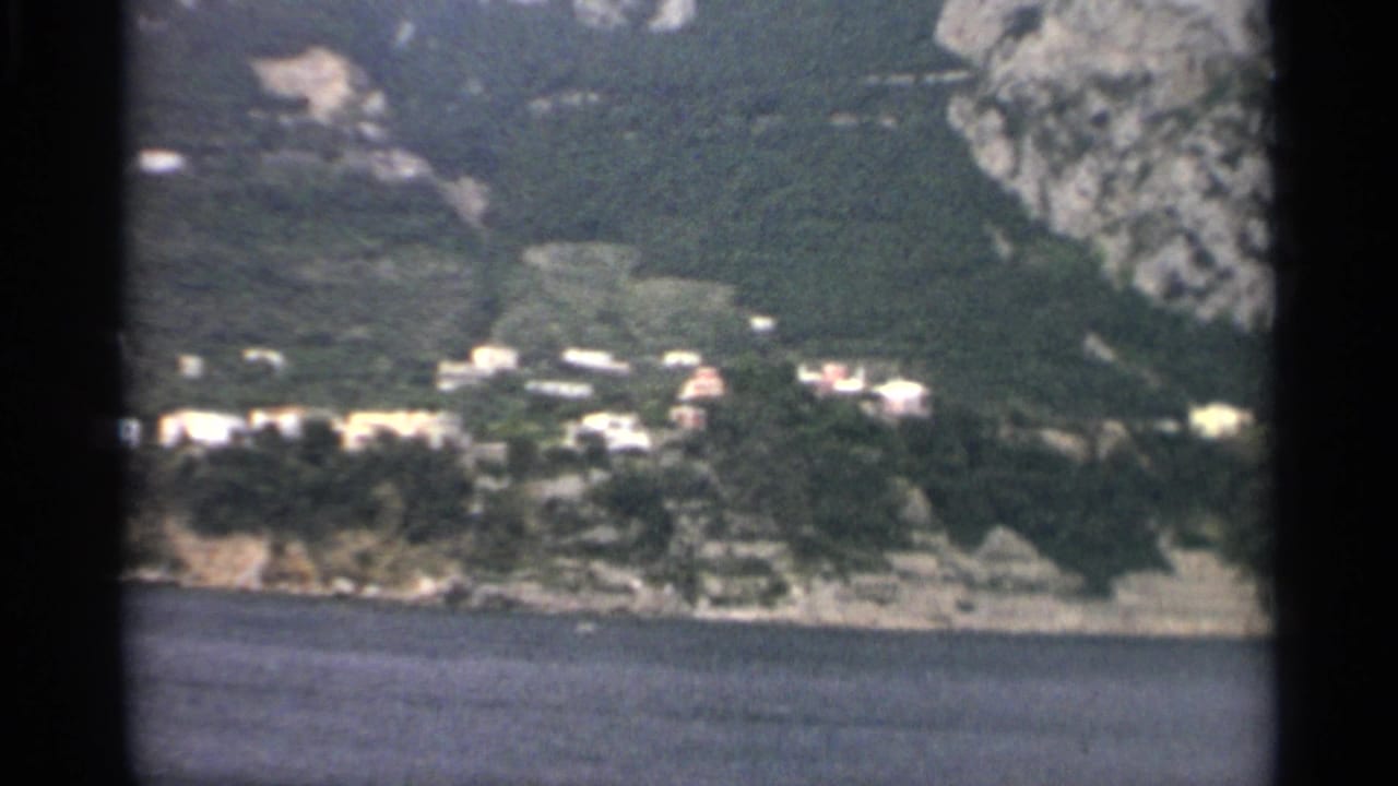 1965 8mm frame showing small shoreline buildings, dense trees, blue water, and a rocky hillside rising behind the settlement.