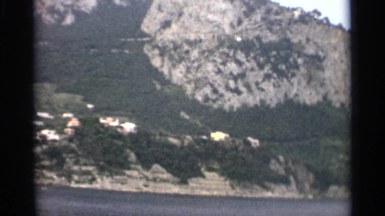 1965 8mm frame with a pale rocky slope, scattered light-colored houses, dark trees, and a narrow shoreline above the water.