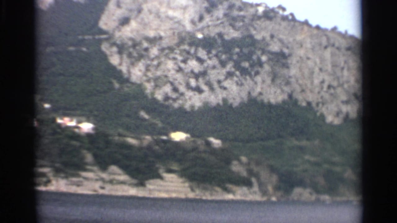 Rocky wooded hillside above calm sea with small pale buildings near the shore.