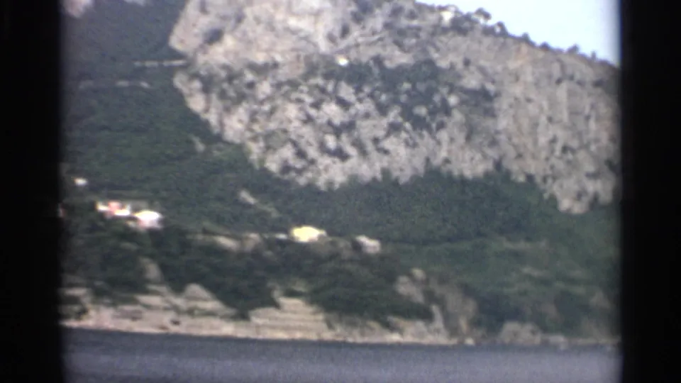1965 Stockholm home-movie frame showing a rocky green hillside, calm sea, and distant pale buildings along the shoreline.