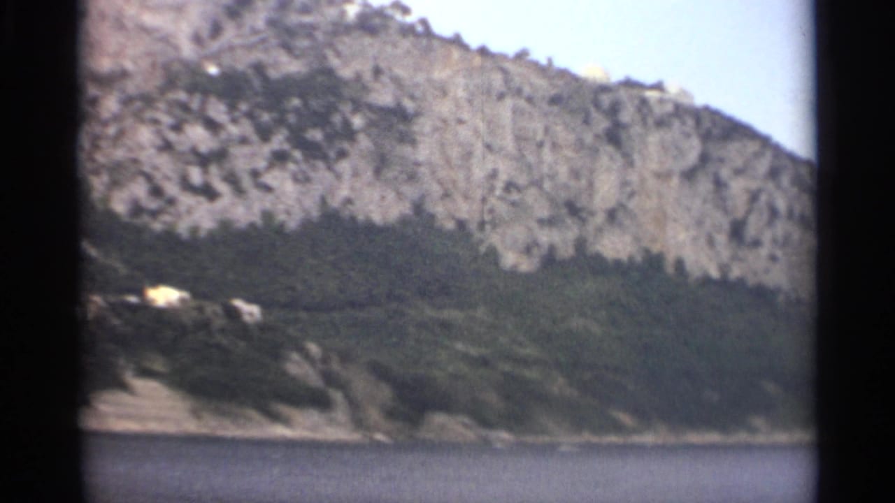 1965 Stockholm coastal view with a rocky wooded slope, calm sea in the foreground, and a few distant hillside buildings.