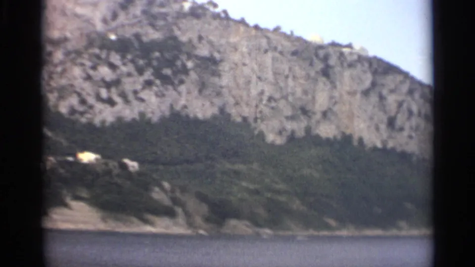 1965 Stockholm coastal view with a rocky wooded slope, calm sea in the foreground, and a few distant hillside buildings.