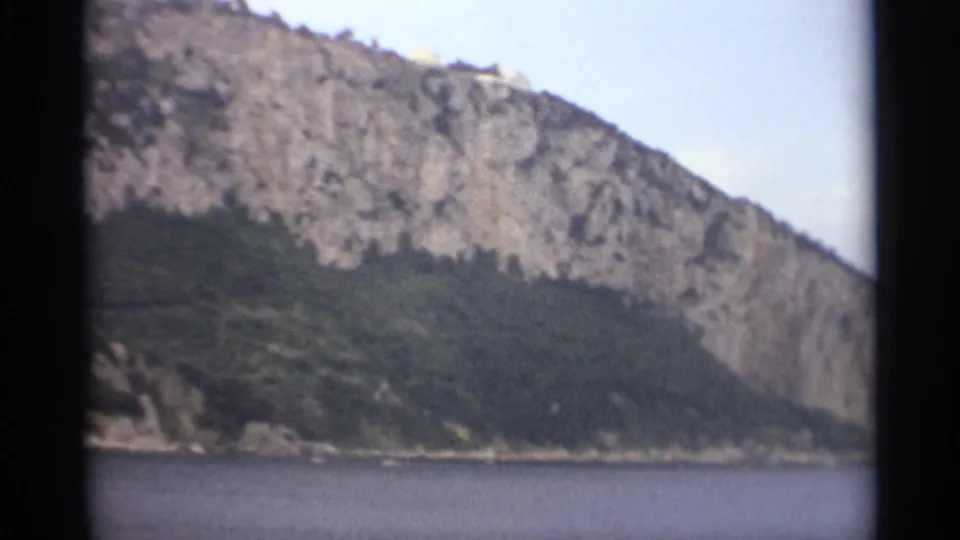 1965 Stockholm 8mm frame showing a steep rock face, dense green vegetation, and calm coastal water below.