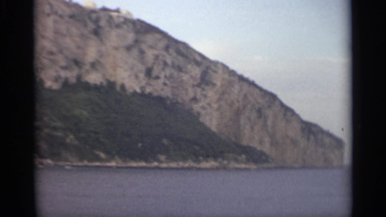 1965 8mm Stockholm travel view of a steep rocky waterfront cliff with dark vegetation below and calm water in the foreground.