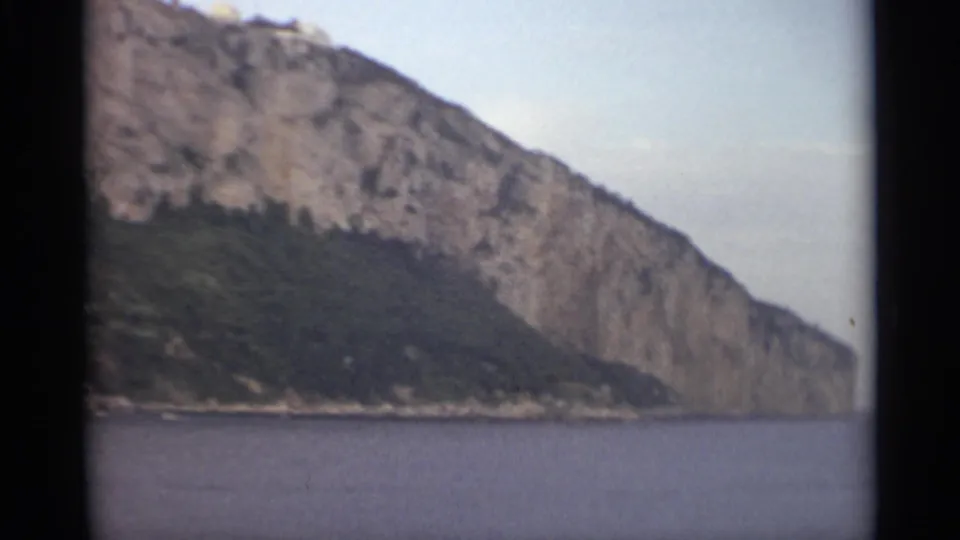 1965 8mm Stockholm travel view of a steep rocky waterfront cliff with dark vegetation below and calm water in the foreground.