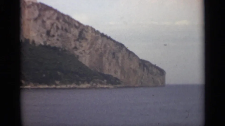 1965 8mm view near Stockholm showing a broad cliff face, low green shoreline, and open water under a hazy sky.