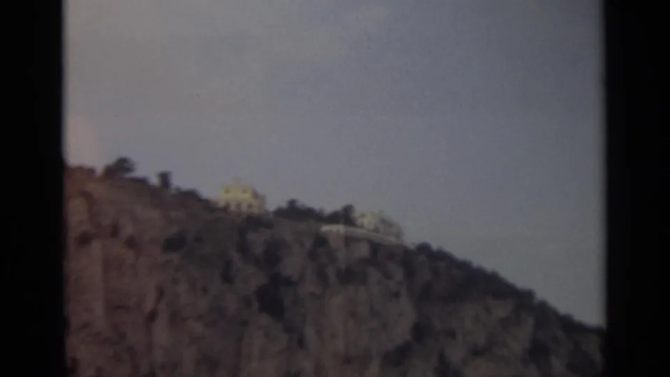 1965 8mm Stockholm view of distant pale buildings perched on a rocky hilltop above the waterfront, seen through soft home-movie focus.