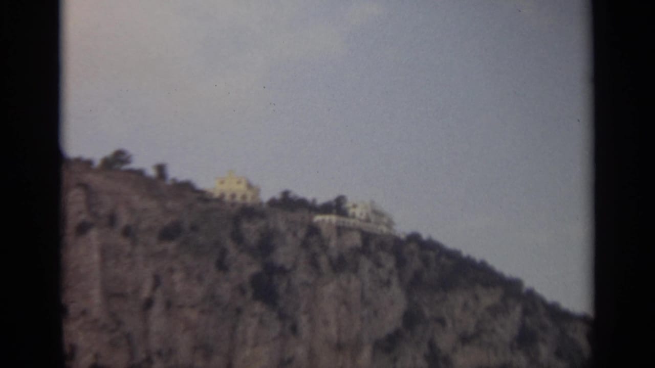 1965 8mm Stockholm view of light-colored buildings perched above a rugged rocky hillside beneath a pale sky.