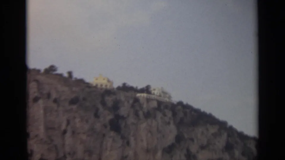 1965 8mm Stockholm view of light-colored buildings perched above a rugged rocky hillside beneath a pale sky.