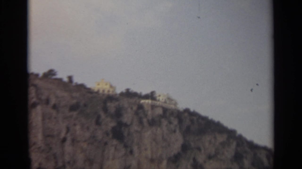 Light-colored buildings sit on top of a rocky cliff with sparse trees and open sky.