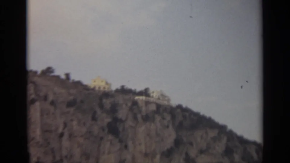 1965 Stockholm home-movie frame showing a rocky cliffside with pale buildings on the ridge and sparse vegetation.