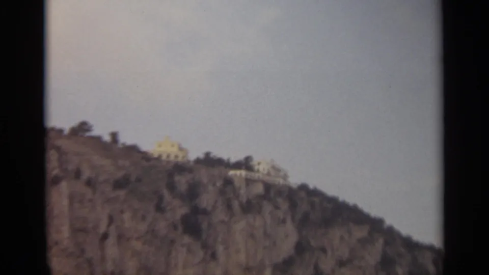 Soft 8mm Stockholm frame of a rocky hillside and distant light-colored buildings under a washed pale sky.