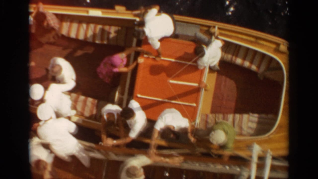 1965 Stockholm home-movie frame showing passengers clustered in a yellow wooden boat with striped seating and bright sun.