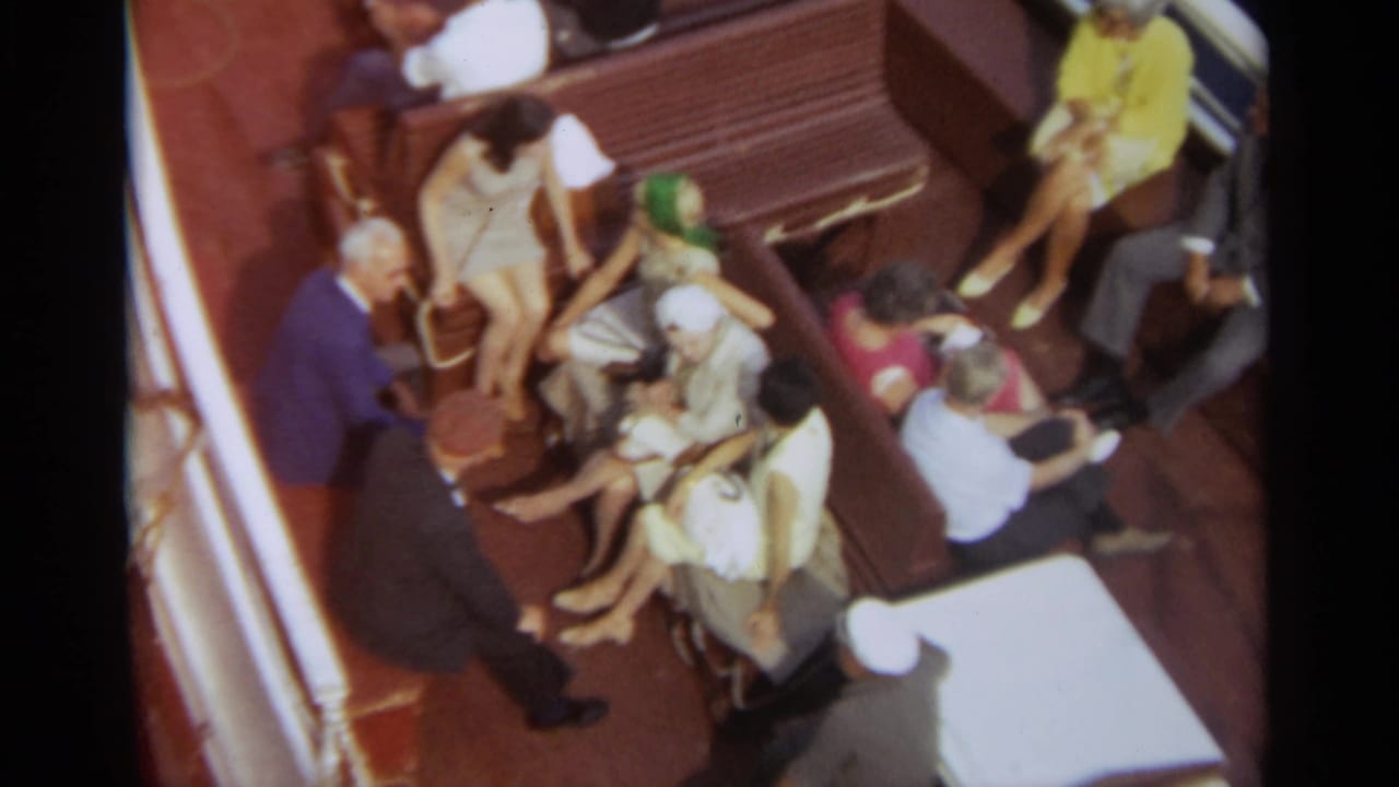 1965 home-movie view of passengers seated on a Stockholm boat deck with wooden benches and blue water nearby.