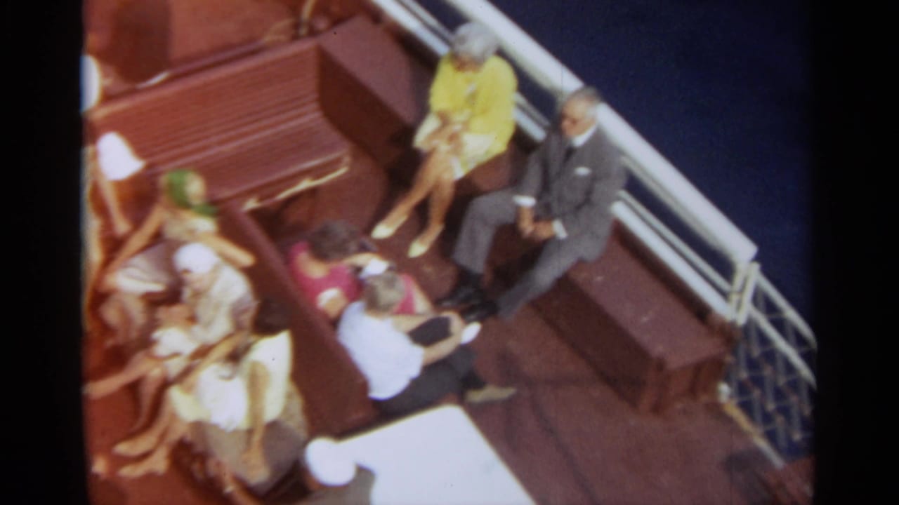 Passengers in suits and dresses sit along benches on a boat deck over blue water.