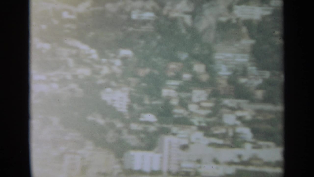 1965 8mm aerial view over Stockholm, with hazy hillside buildings, trees, and a bright shoreline area softened by exposure and grain.