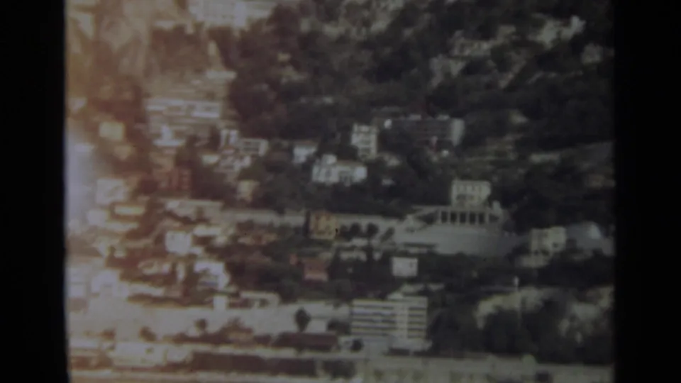 Aerial home-movie frame of Stockholm showing wooded slopes, waterfront structures, shoreline roads, and warm sunlight flare across the left side.