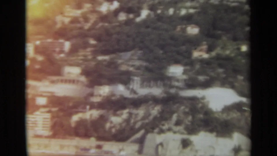 1965 Stockholm aerial frame with clustered hillside buildings, dark tree cover, bright shoreline glare, and soft 8mm color fading.