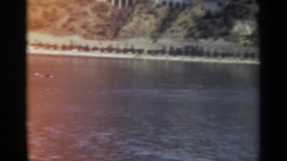 Aerial view of Stockholm shoreline and open water, with a wooded slope, pale coast road, small marks on the water, and strong sun reflection.