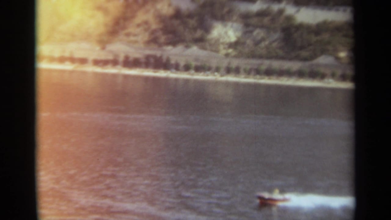 1965 Stockholm 8mm frame showing a small speedboat moving across a broad river with a tree-lined bank behind it.