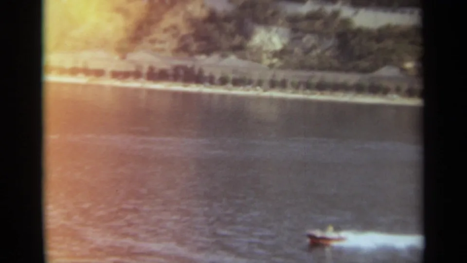 1965 Stockholm 8mm frame showing a small speedboat moving across a broad river with a tree-lined bank behind it.