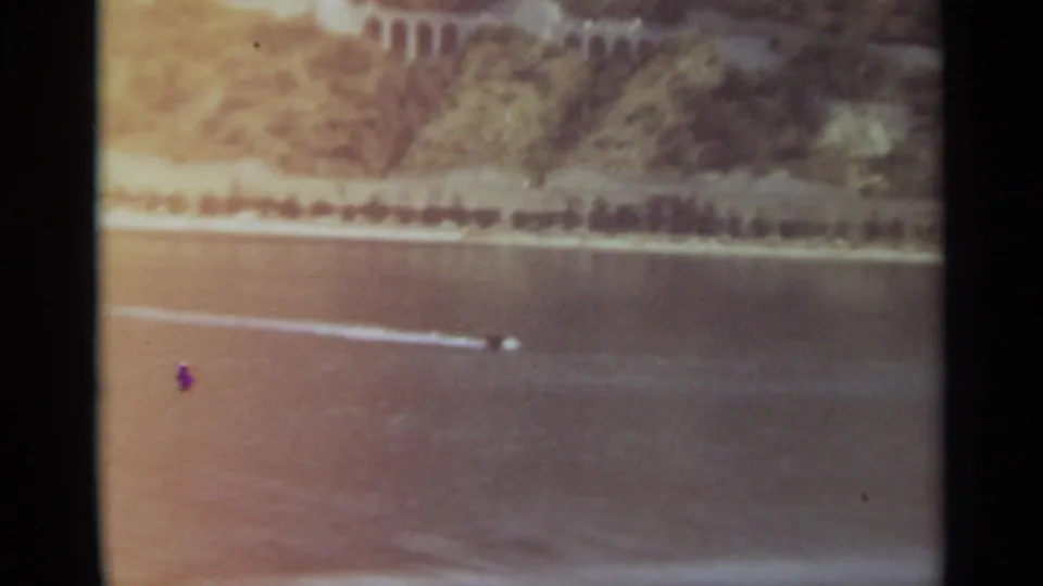 Small speedboat with a bright wake passes across a Stockholm river, with wooded hillside and shoreline visible beyond.