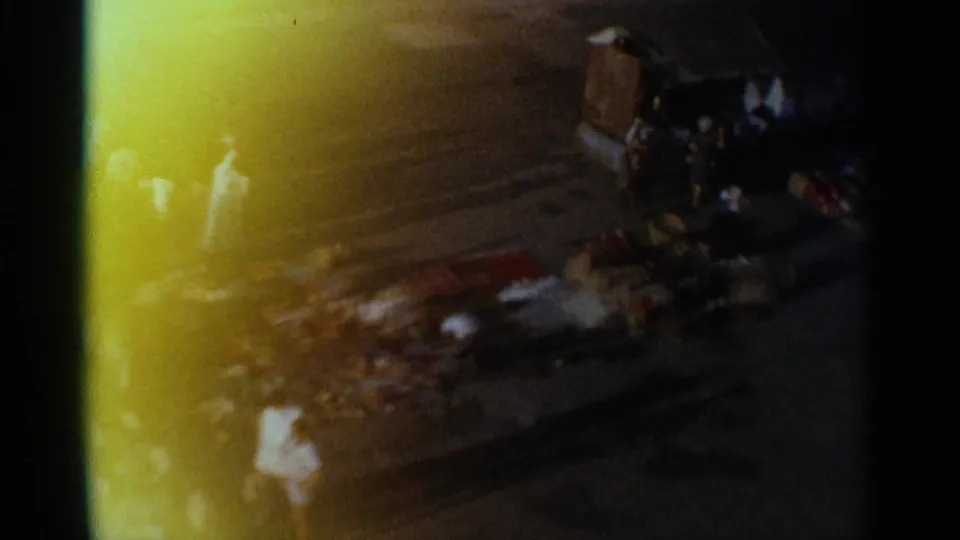 Handheld 8mm view of a Tangier street-market area, with pedestrians, scattered goods, and a person in white partly obscured by glare.