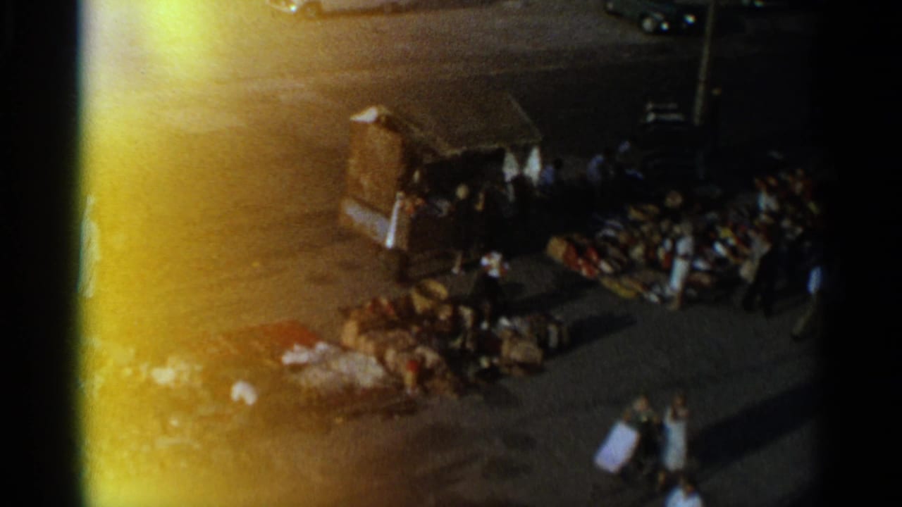 High-angle market scene with a kiosk, shoppers and piles of goods on pavement.